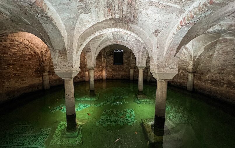 Flooded fifth-century crypt of Basilica di San Francesco; green water and goldfish glide over ancient floor mosaics between brick arches.