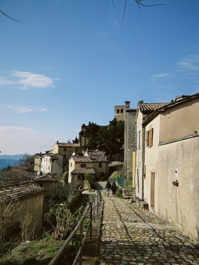 Cobbled street in San Leo lined with stone houses under a blue sky.