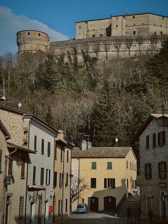 View of the San Leo fortress rising above the village houses, with stone walls and towers set against a wooded hillside.
