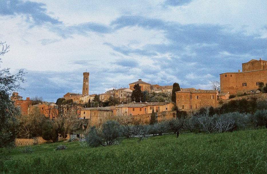 Distant view of Santarcangelo di Romagna at dusk, with its medieval clock tower and stone buildings surrounded by fields and olive trees.