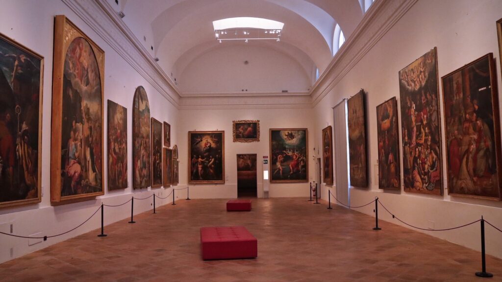 Faenza Pinacoteca art gallery hall with classical paintings, high vaulted ceiling, and red museum benches