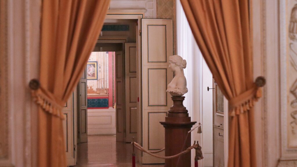 Marble bust framed by rust-colored curtains in a Palazzo Milzetti corridor with classical Italian decor