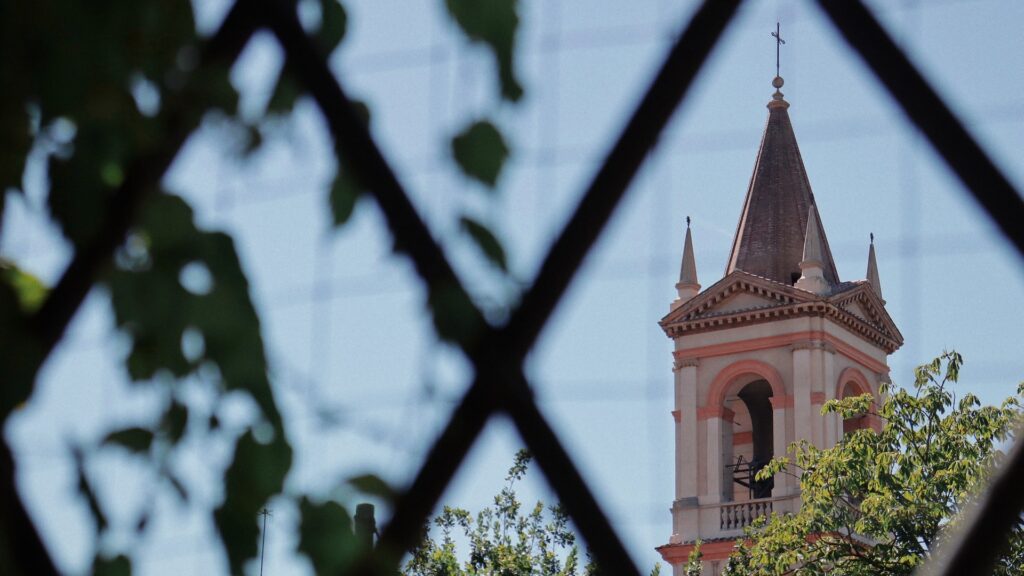 Church bell tower seen through leafy branches and a wrought iron fence in Faenza