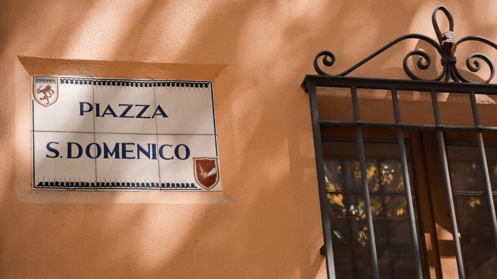 Piazza S. Domenico ceramic street sign on a warm stucco wall beside an iron window grille