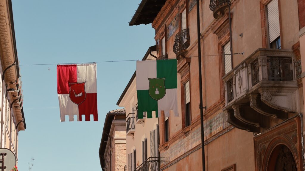 Historic street in Faenza with hanging district flags, brick architecture, balconies, and blue summer sky
