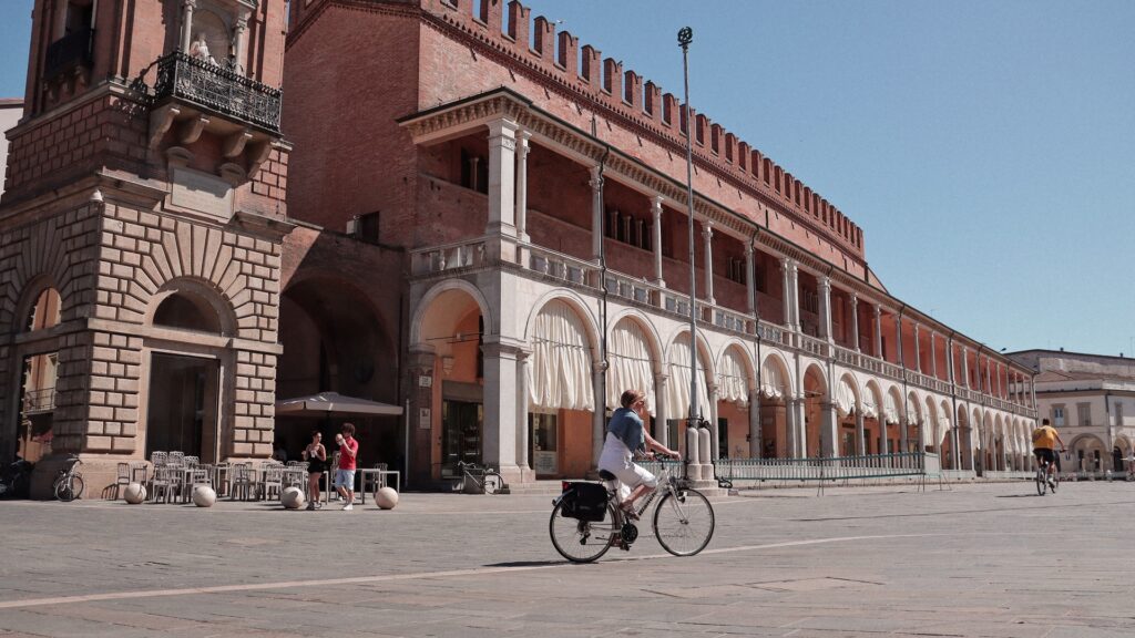 Cyclist passing an arcaded brick palace in a spacious Piazza del Popolo in Faenza under clear blue sky