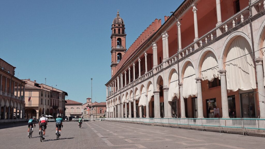 Cyclists riding through a wide Piazza del Popolo in Faenza lined with arcades, columns, and a bell tower