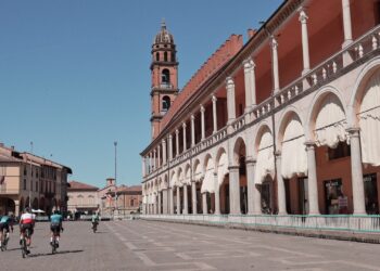 Cyclists riding through a wide Piazza del Popolo in Faenza lined with arcades, columns, and a bell tower