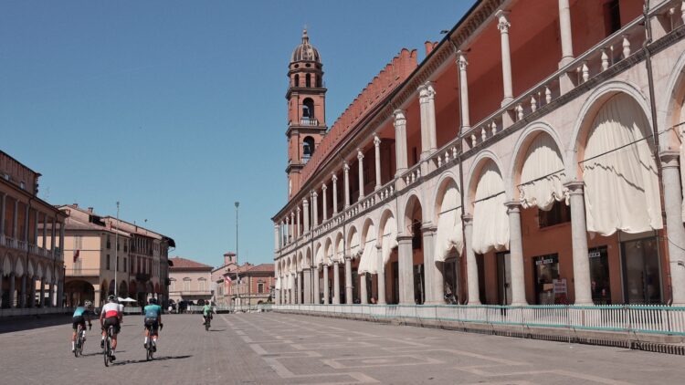 Cyclists riding through a wide Piazza del Popolo in Faenza lined with arcades, columns, and a bell tower